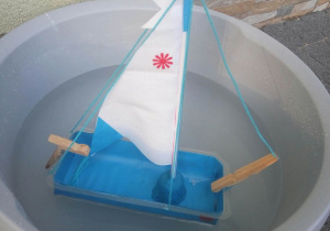 Biało- niebieska łódka wykonana z papieru, pudełka plastikowego i sznurka kołysząca się na wodzie w miseczce